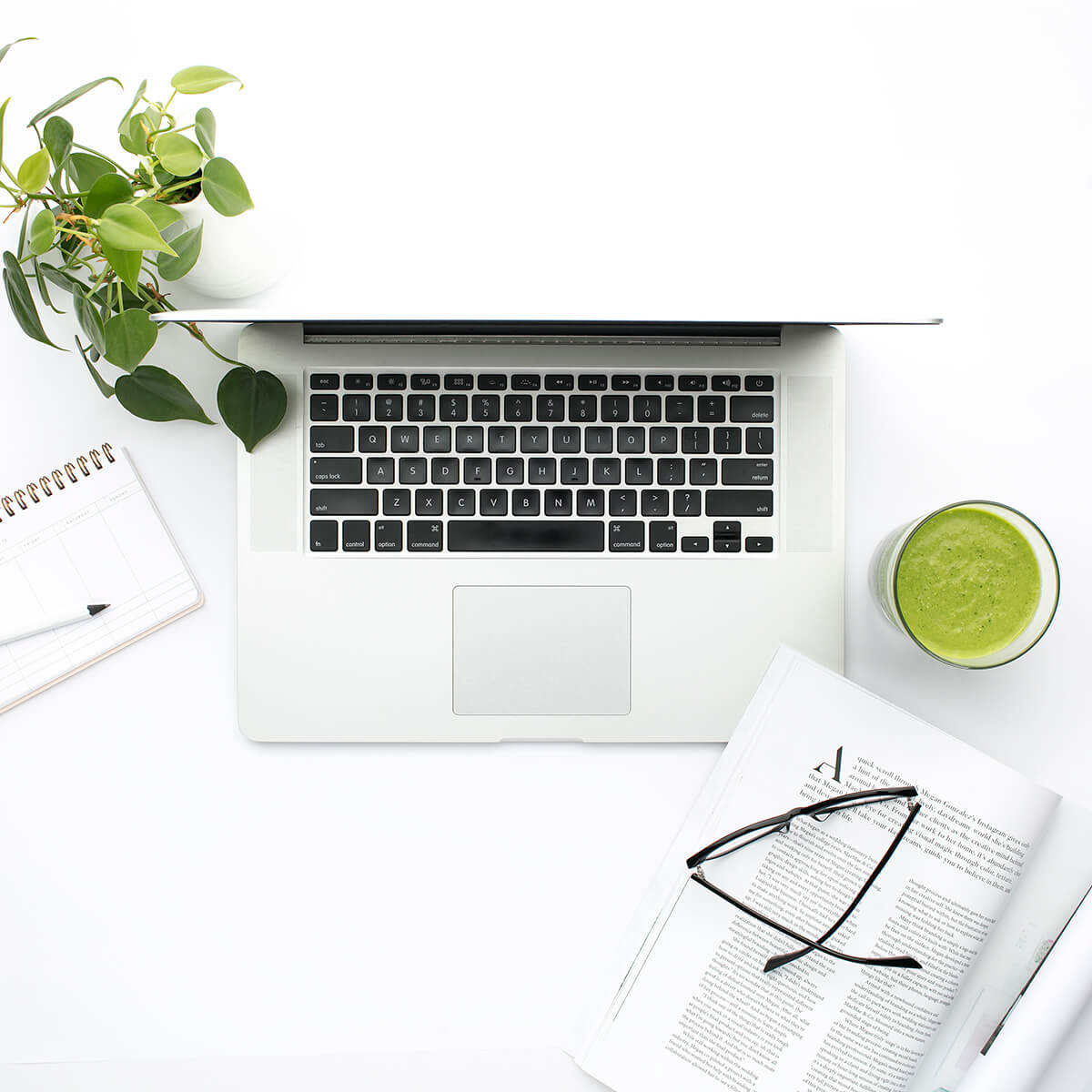 Laptop and book on a white desk with green plant and green milkshake